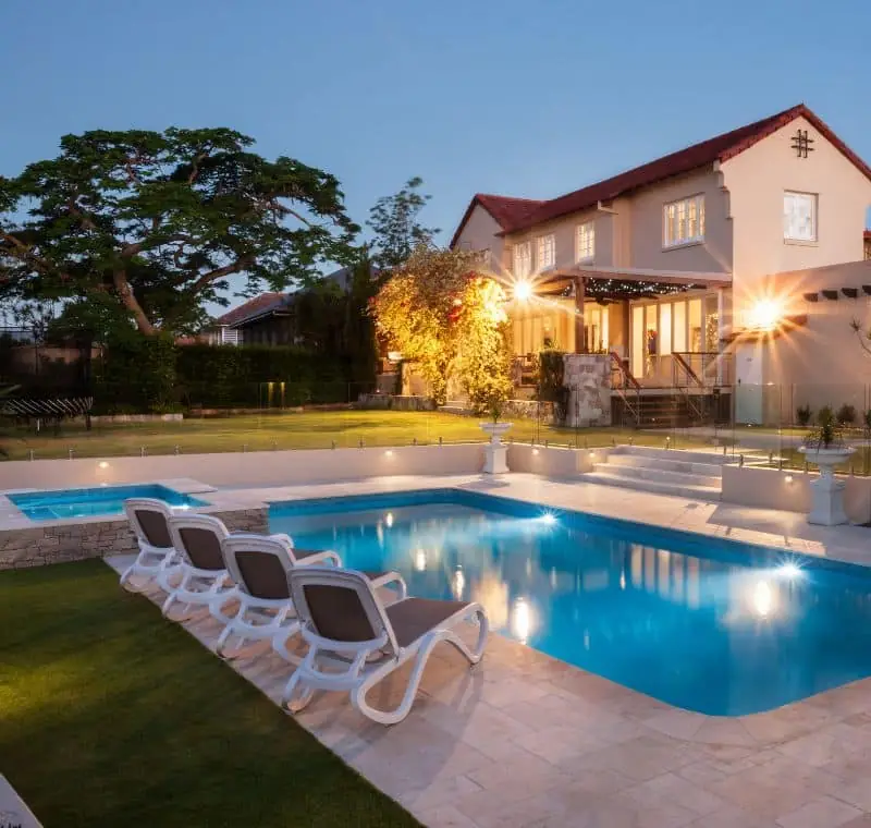 Pool with loungers on a lawn beside a stucco home lit at dusk