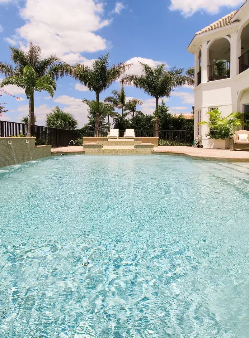 Wide shallow pool with clear water behind a two-story home and palm trees