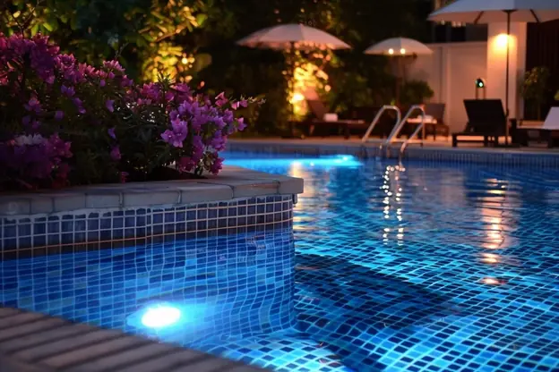 Mosaic-tile pool lit at night with flowering planters and umbrellas